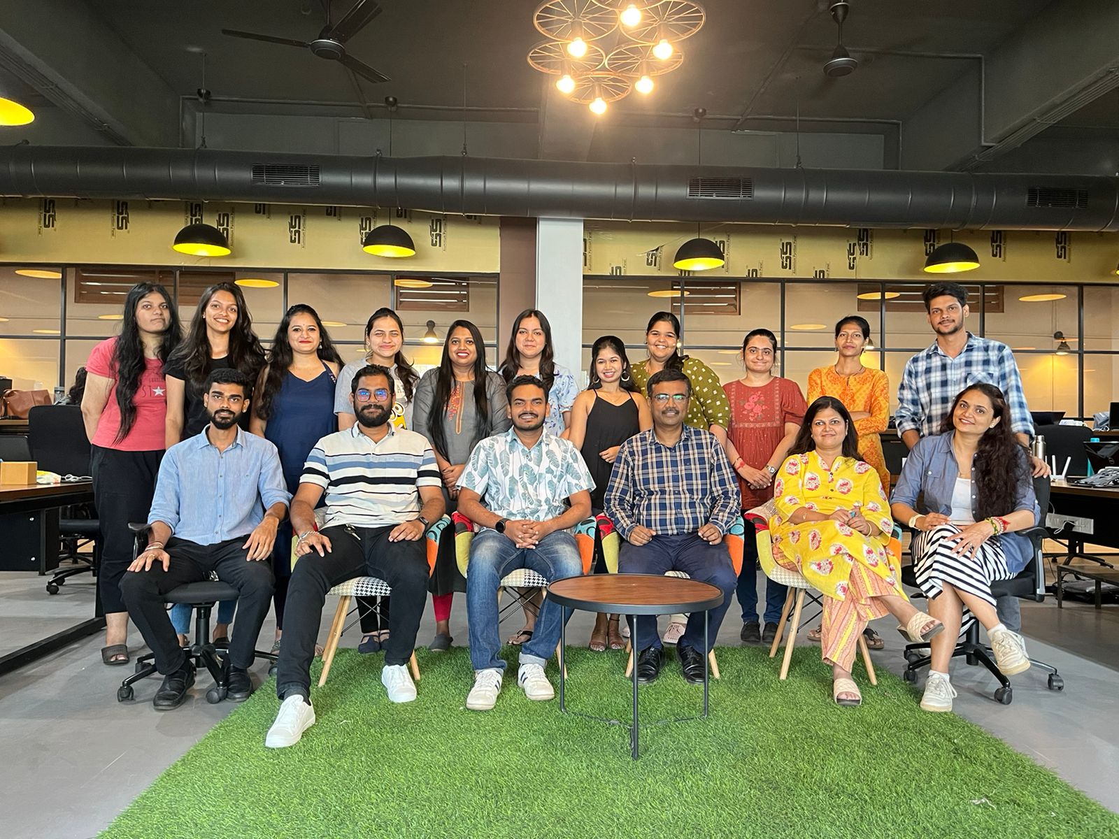 Group of 15 colleagues posing for a group photo in a modern office with a green carpet and glass-walled background.