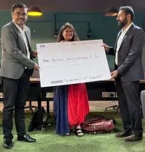Three people, two men in suits and a woman in colorful attire, hold a large ceremonial check outdoors during a presentation.
